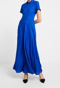 Sininen nappikauluspaita, jossa on lyhyet hihat, istuva vyötärö, virtaava hame ja sileä tekstuuri. Yhdistetty mustiin kärkitossuihin.