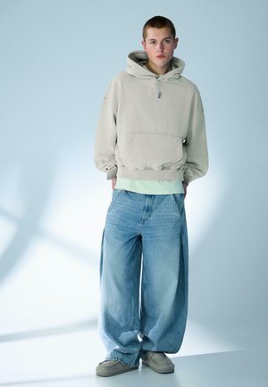 Mladý človek stojí s rukami v kapsách, oblečený v béžovej mikine s kapucňou, svetlozelenom tričku, modrých nohaviciach so širokými nohavicami a sivých teniskách na bledom pozadí.