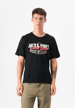 Young man wearing a black graphic t-shirt with "Jack & Jones" printed on it, standing against a plain white background.