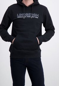 Homme portant un sweat à capuche noir avec le texte "LINDBERGH Copenhagen. Paris. New York", les mains dans la poche avant, assorti d'un pantalon noir.