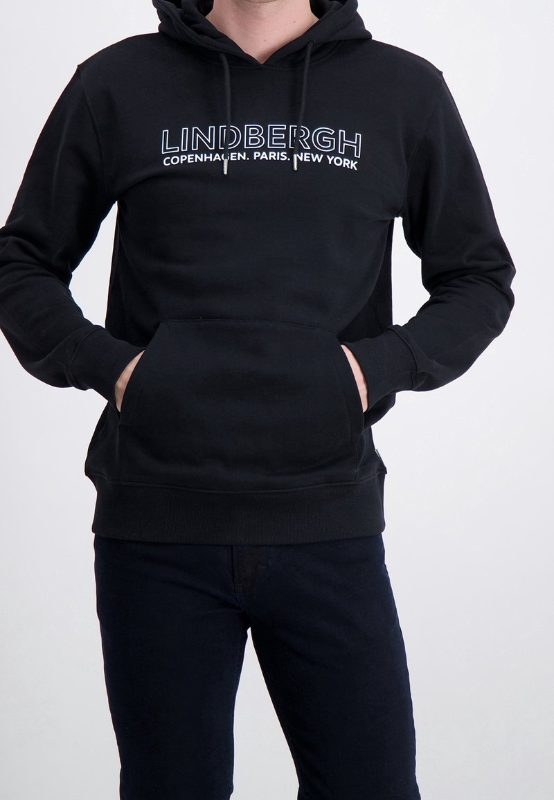 Homme portant un sweat à capuche noir avec le texte "LINDBERGH Copenhagen. Paris. New York", les mains dans la poche avant, assorti d'un pantalon noir.