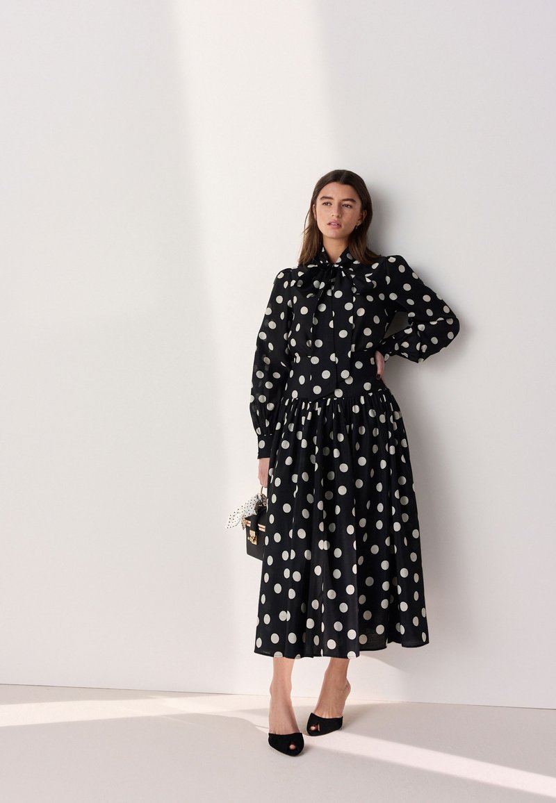 Femme debout contre un mur blanc portant une robe midi noire à manches longues avec de grands pois blancs, tenant un sac à main noir, portant des talons noirs.