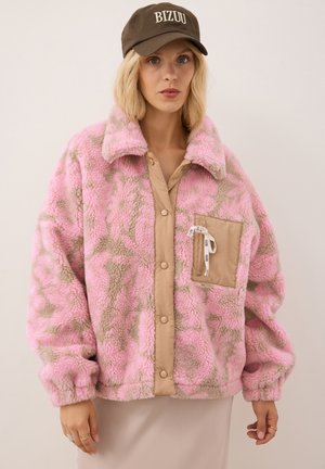 Vrouw draagt een roze en beige fleecejack met patroon, met een beige knopenbies en zak, en een donkerbruine pet met opschrift "BIZUU."