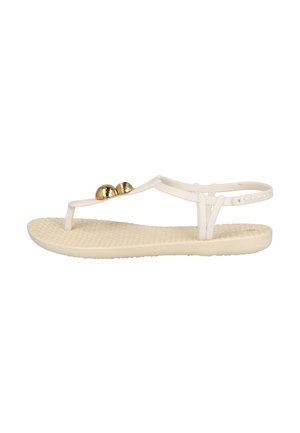 Sandalia plana beige con suela texturizada, cuentas decorativas doradas en la tira entre los dedos y cierre ajustable en el tobillo con botón.