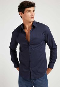 Camisa de manga larga azul marino oscura, confeccionada en tejido suave, que cuenta con un cierre frontal de botones, cuello y un discreto detalle de logo en el pecho.