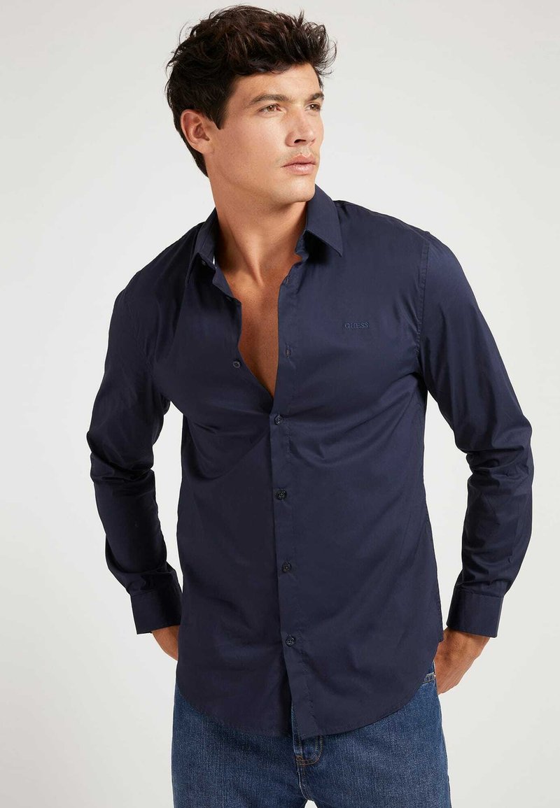 Camisa de manga larga azul marino oscura, confeccionada en tejido suave, que cuenta con un cierre frontal de botones, cuello y un discreto detalle de logo en el pecho.