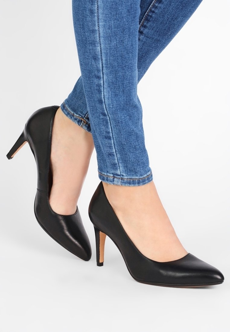 Pump in pelle nera con tacco alto a stiletto e punta affusolata, abbinati a jeans blu attillati. Tessuto liscio, senza motivi visibili.