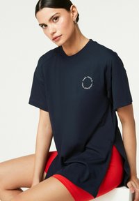 Tricou din bumbac de culoare albastru marin, cu mâneci scurte și decolteu rotund, având un logo circular alb pe piept. Şorturi roșii vizibile dedesubt.