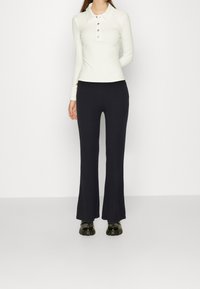 Witte poloshirt met lange mouwen en kraag, voorzien van vijf knopen, gecombineerd met zwarte getailleerde flared broek. Model draagt zwarte chunky sneakers.