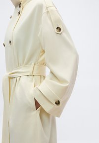 Κρεμ παλτό τύπου trench με κουμπωτές επιχρίσεις στους ώμους και τις μανσέτες, ζωνάρι στη μέση και πλαϊνή τσέπη με το χέρι εν μέρει μέσα.