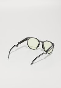 Transparente graue Sonnenbrille mit eckigen Rahmen, grün getönten Gläsern und einzigartig geformten Bügeln. Auffälliges Zickzack-Detail an den Schläfen.