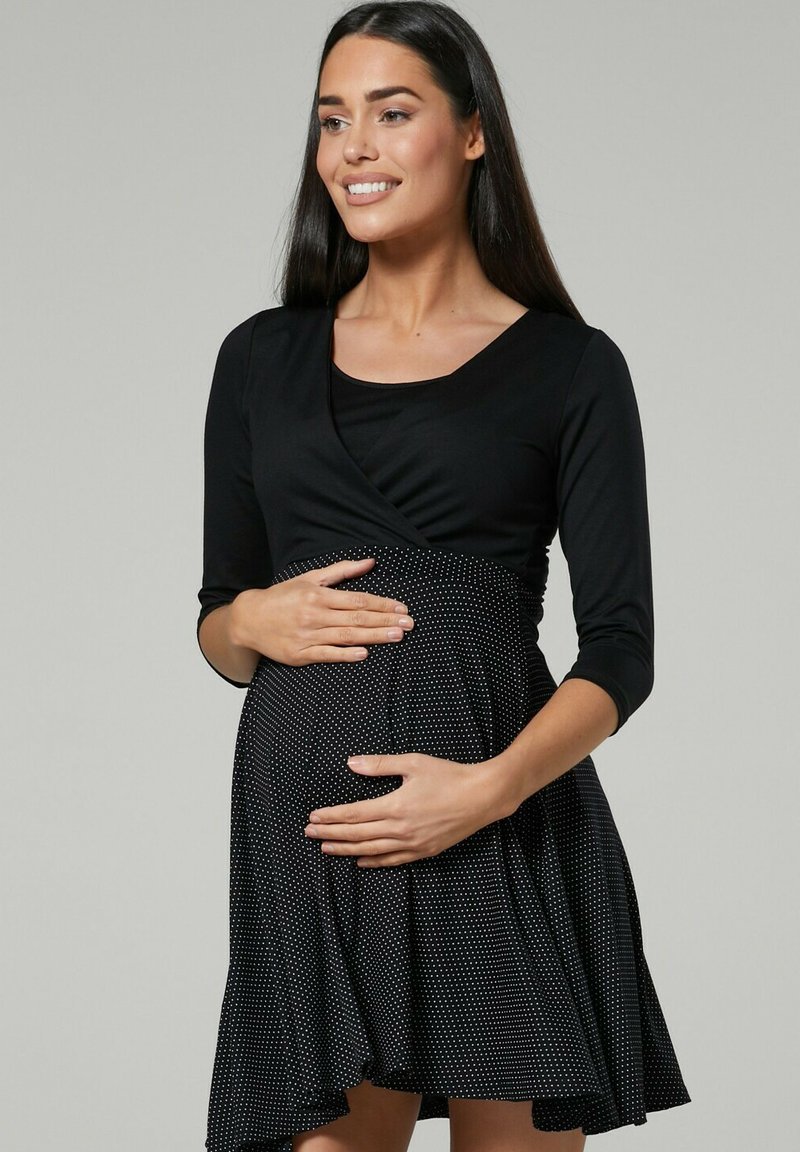 Black maternity dress with a wrap design. Long sleeves, dotted skirt, and a smooth texture. The model holds her belly.