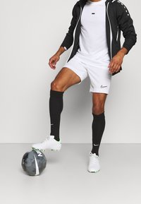 Svart zip-up jacka med vita detaljer, vit skjorta, vita shorts, svarta strumpor och vita fotbollsskor med gröna detaljer, stående på en grå fotboll.