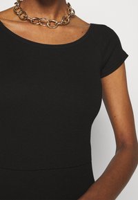 Svart ribbad topp med en figurnära design, korta ärmar och en bred halsringning, kompletterad med ett kraftigt halsband i guld.