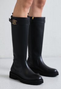 Bottes en caoutchouc noires montant jusqu'aux genoux avec une semelle épaisse, présentant une bande dorée en haut et une finition lisse mate.