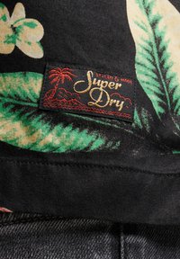 Tessuto nero con una stampa di foglie tropicali in verde e beige. Presenta un'etichetta "Super Dry" rossa e dorata con illustrazione di palma.