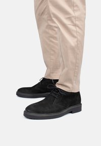 Botas de tobillo de ante negro con puntera redonda, cordones y suela de goma plana, combinadas con pantalones slim-fit de color beige claro.