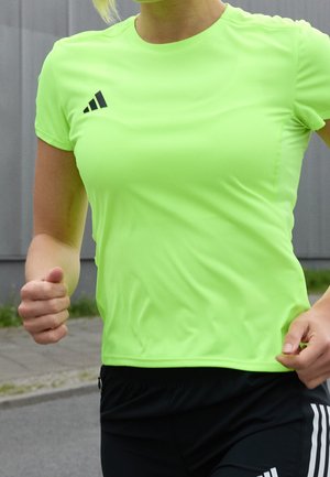 Person läuft im Freien und trägt ein leuchtend neongrünes Adidas-Shirt und schwarze Shorts mit weißen Streifen.