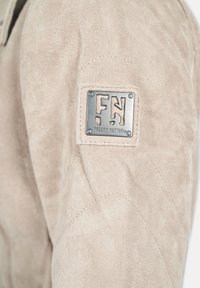 Manche de veste en daim beige avec un patch métallique cousu, présentant des lettres découpées "F N" et "Freaky Nation" gravées en dessous.