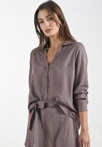 Camicia a maniche lunghe in morbido tessuto testurizzato con sfondo marrone e motivo a piccoli pois bianchi. Presenta un colletto e un legaccio in vita.