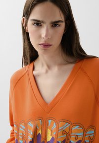 Sweatshirt oversize orange à encolure en V avec un graphique audacieux "REAL" mettant en avant des accents multicolores et une finition en tissu texturé.