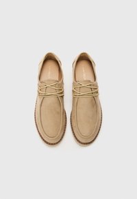 Beige Wildlederschuhe mit runder Zehenform, ausgestattet mit hellen Schnürsenkeln und goldenen Hardware-Akzenten sowie einer kontrastierenden braunen Sohle. Elegantes und minimalistisches Design.