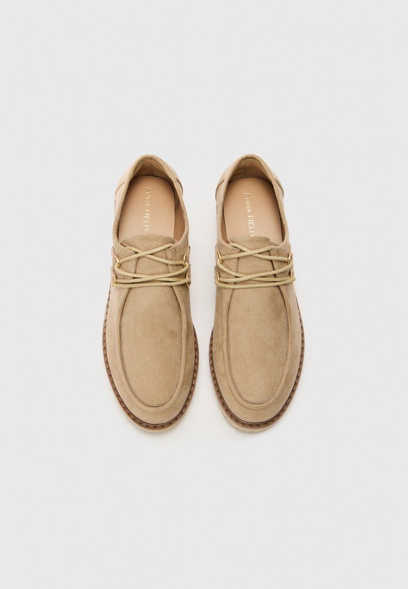 Scarpe in suede beige con punta arrotondata, dotate di lacci chiari e dettagli in hardware dorato, e una suola marrone a contrasto. Design elegante e minimale.