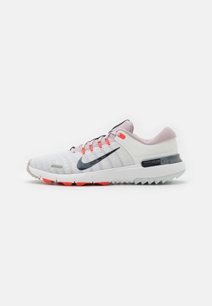 Chaussure de course Nike gris clair et rose avec le swoosh noir, œillets orange et semelle blanche à crampons rouges, vue de profil sur fond blanc.