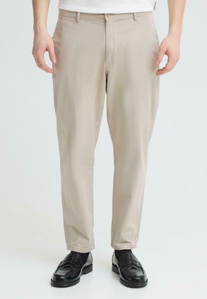 Man draagt beige rechte broek, zwarte leren loafers en witte sokken, staand tegen een effen lichte achtergrond.