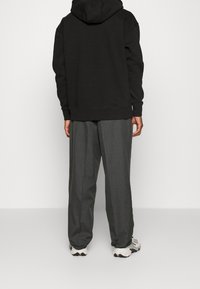 Persona con sudadera negra, pantalones anchos grises y zapatillas blancas de pie, de espaldas, contra un fondo claro y liso.