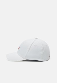 Hvit baseballcap laget av bomull. Har en buet skjerm, seks-panel design, og en liten rød logo-patch på fronten. Glatt tekstur.
