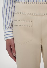 Pantalon beige avec des détails en clous argentés sur la poche et la ceinture, associé à un poignet de chemise à manches longues rayé bleu et blanc.