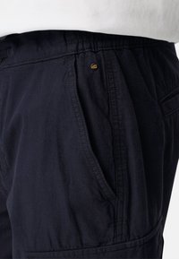 Gros plan sur un pantalon noir à taille élastique avec poche latérale et petit logo en métal rond près de la poche.