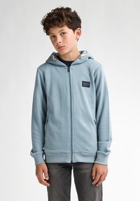 Hooded zip-jacka i ljusblått tyg med sidofickor och en logotypapplikation. Muddar och nederkant med ribbad textur, kragfoder med kontrasterande mönster.