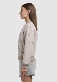 Licht taupe bomberjack met een zachte textuur, geribbelde boorden en tailleband, zijzakken en een logo-opdruk op de linkermouw.