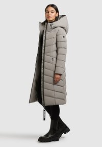 Langer grauer Daunenmantel mit Reißverschluss, weitem Kapuzen und gestepptem Muster. Über einem schwarzen Outfit und klobigen schwarzen Stiefeln getragen.