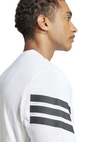 Camisa blanca de manga larga con tres rayas horizontales negras en el hombro izquierdo, hecha de una tela suave con una textura lisa.