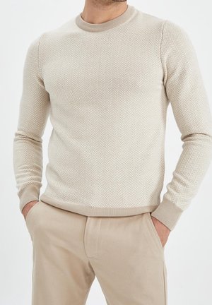 Mann trägt beige Fischgrät-Strickjacke und beige Hose mit Händen in den Taschen, vor einfachem weißem Hintergrund.