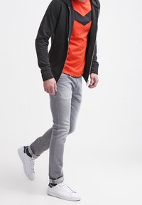 Černá mikina s kapucí na zip přes oranžové tričko s chevron vzorem, doplněná o světle šedé džíny a bílé tenisky s černými akcenty.