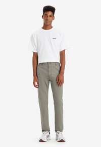 Camiseta blanca de manga corta con logo negro, combinada con pantalones verde oliva claros. El calzado presenta toques en blanco y negro. Fondo liso.