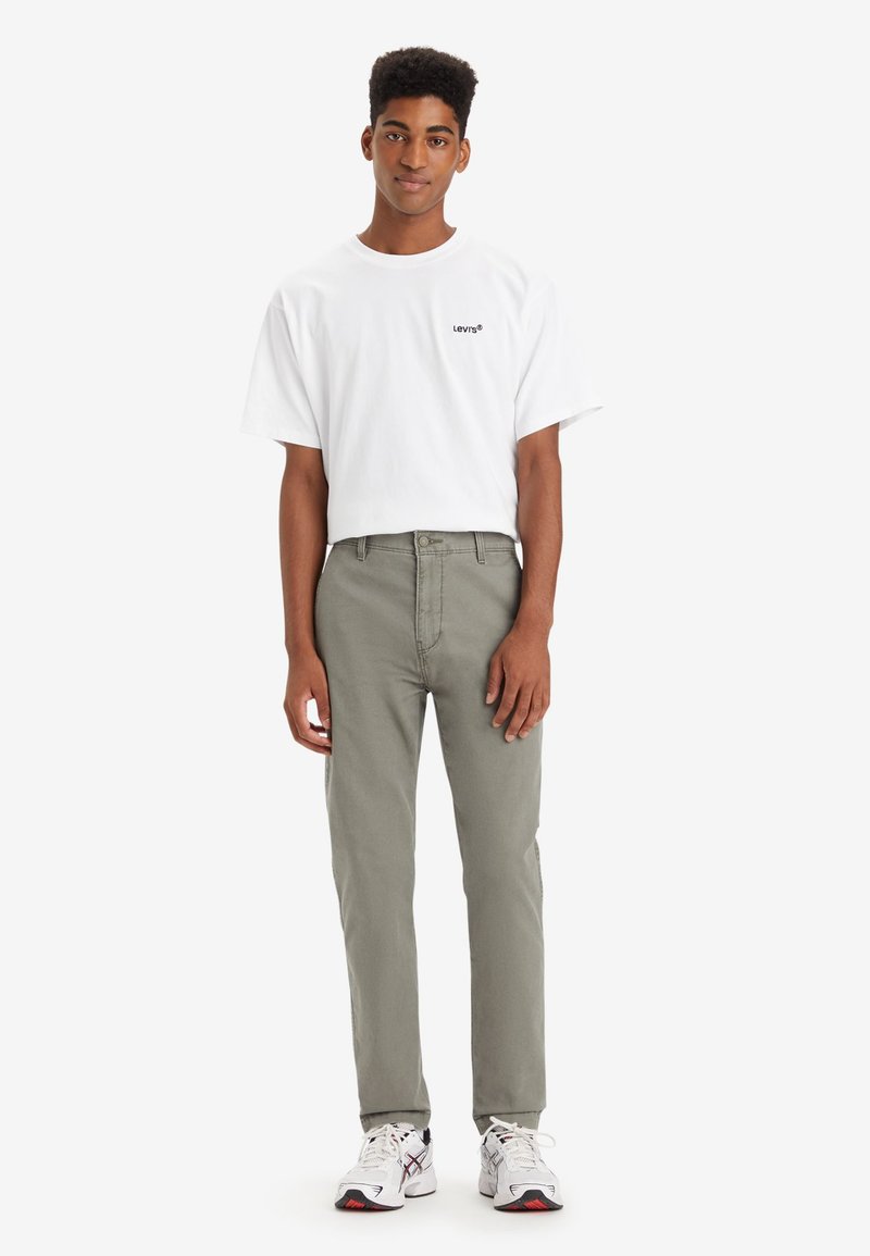 Camiseta blanca de manga corta con logo negro, combinada con pantalones verde oliva claros. El calzado presenta toques en blanco y negro. Fondo liso.
