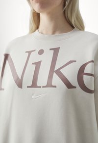 Béžová mikina s velkým vínově červeným nápisem "Nike" a malým bílým logem Nike ve tvaru šípu. Design s kulatým výstřihem a žebrovanou texturou.