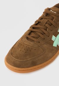 Barna velúr sportcipő lyuggatott orr-résszel, zöld részletekkel és barna fűzővel. Gumi talp, formázott alak és varrott díszítés.