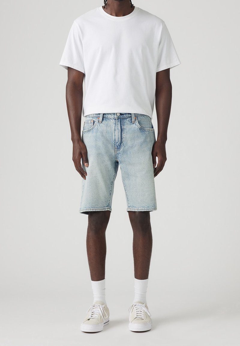 Personne portant un t-shirt blanc uni, un short en denim bleu clair, des chaussettes blanches et des baskets beiges, debout devant un fond gris.