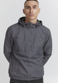 Sivi windbreaker jakna s polu-zipom, podesivom kapuljačom, elastičnim manžetama i prednjim džepom. Ima brendiranu oznaku na prsima.