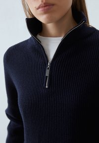 Marineblauer gerippter Pullover mit einem Viertel-Reißverschlusskragen und silbernen Reißverschlussdetails, über einem weißen Hemd getragen und mit strukturierter Stoffoptik.