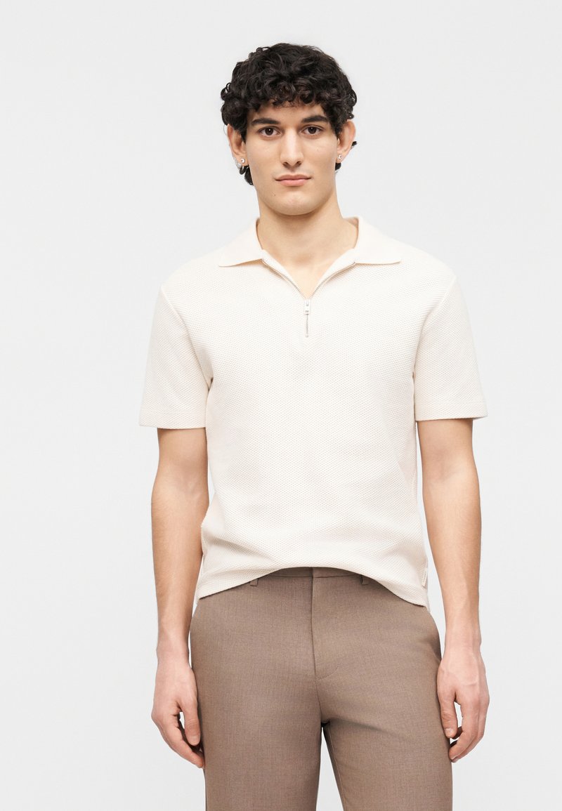 Νέος άνδρας που φοράει μπεζ κοντομάνικο πόλο πουκάμισο με φερμουάρ στον γιακά και καφέ παντελόνι, στέκεται μπροστά σε ένα απλό φωτεινό φόντο.