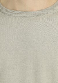 Zbliżenie jasnobeżowej dzianiny swetra, ukazujące okrągły dekolt oraz drobne ryglowanie kołnierza.