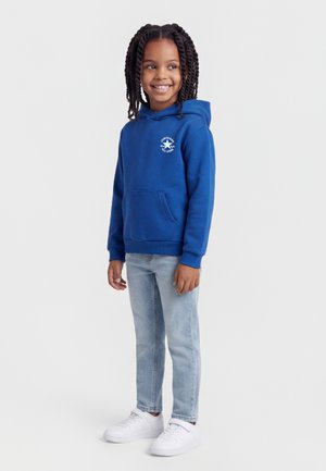 Bambino sorridente con capelli intrecciati che indossa una felpa con cappuccio Converse blu, jeans azzurri e scarpe da ginnastica bianche, in piedi davanti a uno sfondo semplice.