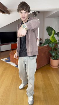 Person wearing a brown zip-up hoodie, black t-shirt, light gray jeans, and silver sneakers, pointing at a gold watch with a green face.